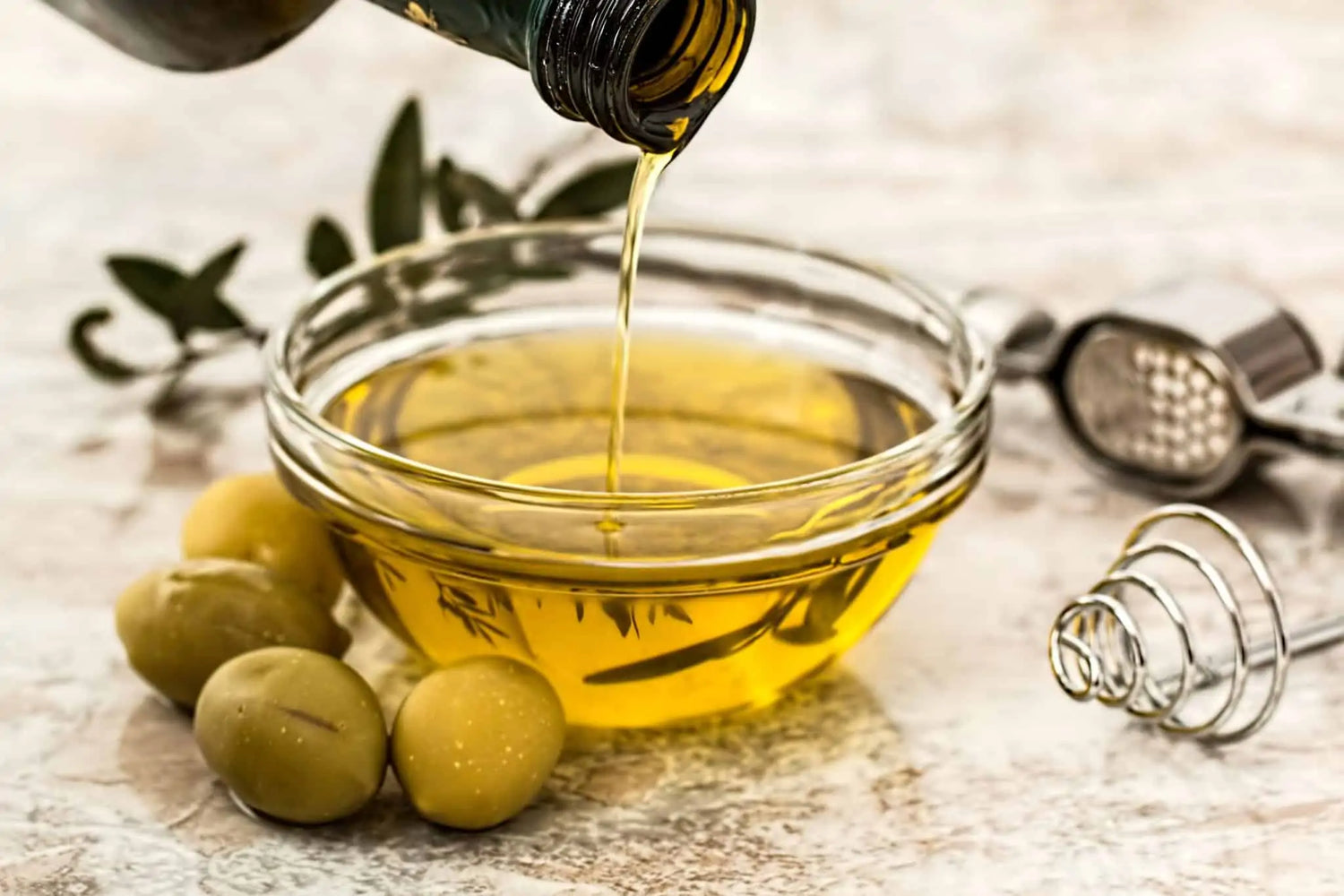 Olive oil being poured into a glass bowl with olives and a grater on a wooden surface.